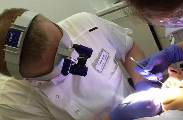 Tannlege Dr. Laszlo Lukacs ved Helvetic Clinics i Budapest utfører en nøyaktig rotbehandling på en pasient, utstyrt med Zeiss lupebriller, mens en assistent bistår med tannlegeinstrumenter.